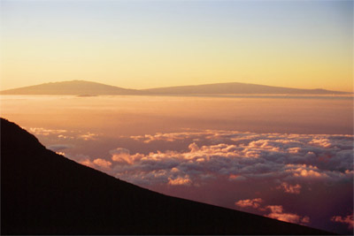 Figure 1.14: Sunrise view of Hawai’i Island from the summit of Haleakalā, Maui. Four of Hawai’i’s five subaerial volcanoes are visible. On the leftmost side is Mauna Kea, in the post-caldera stage, and smaller, eroded Kohala peeks through the clouds directly in front. Mauna Loa, a giant shield volcano is on the right, and on its flank is smaller Hualālai, also a post-caldera volcano. Although the topography appears gentle and subdued, both Mauna Kea and Mauna Loa rise more than 4,000 meters (13,100 feet) above sea level.